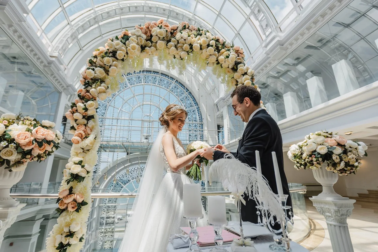 Couple exchanging rings under a flower arch in a glass atrium