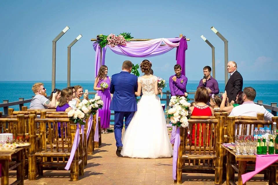 Ceremony on a pier above the sea with a purple-draped arch and guests on benches