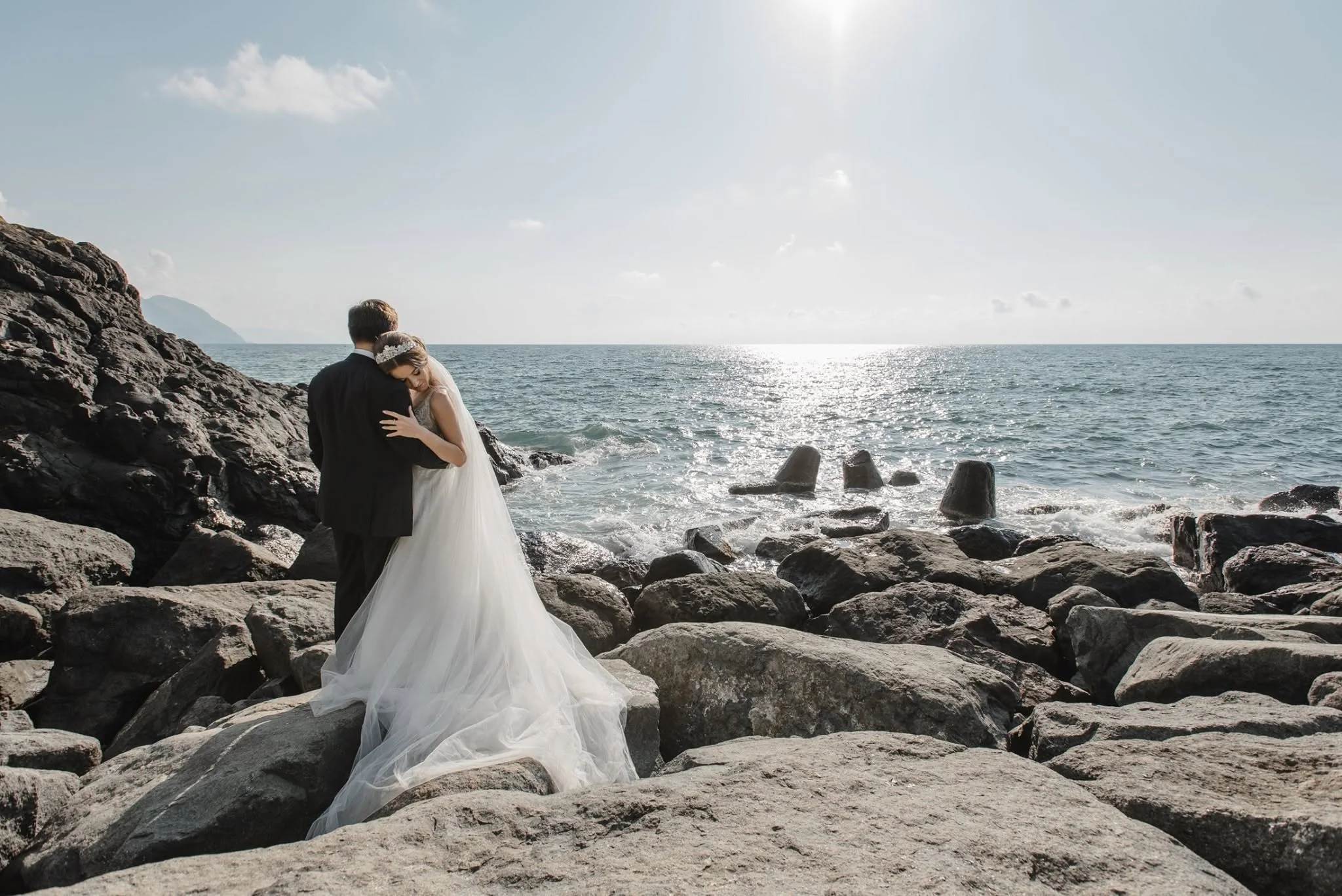Bride and groom embracing on the rocky shore against the sun over the sea