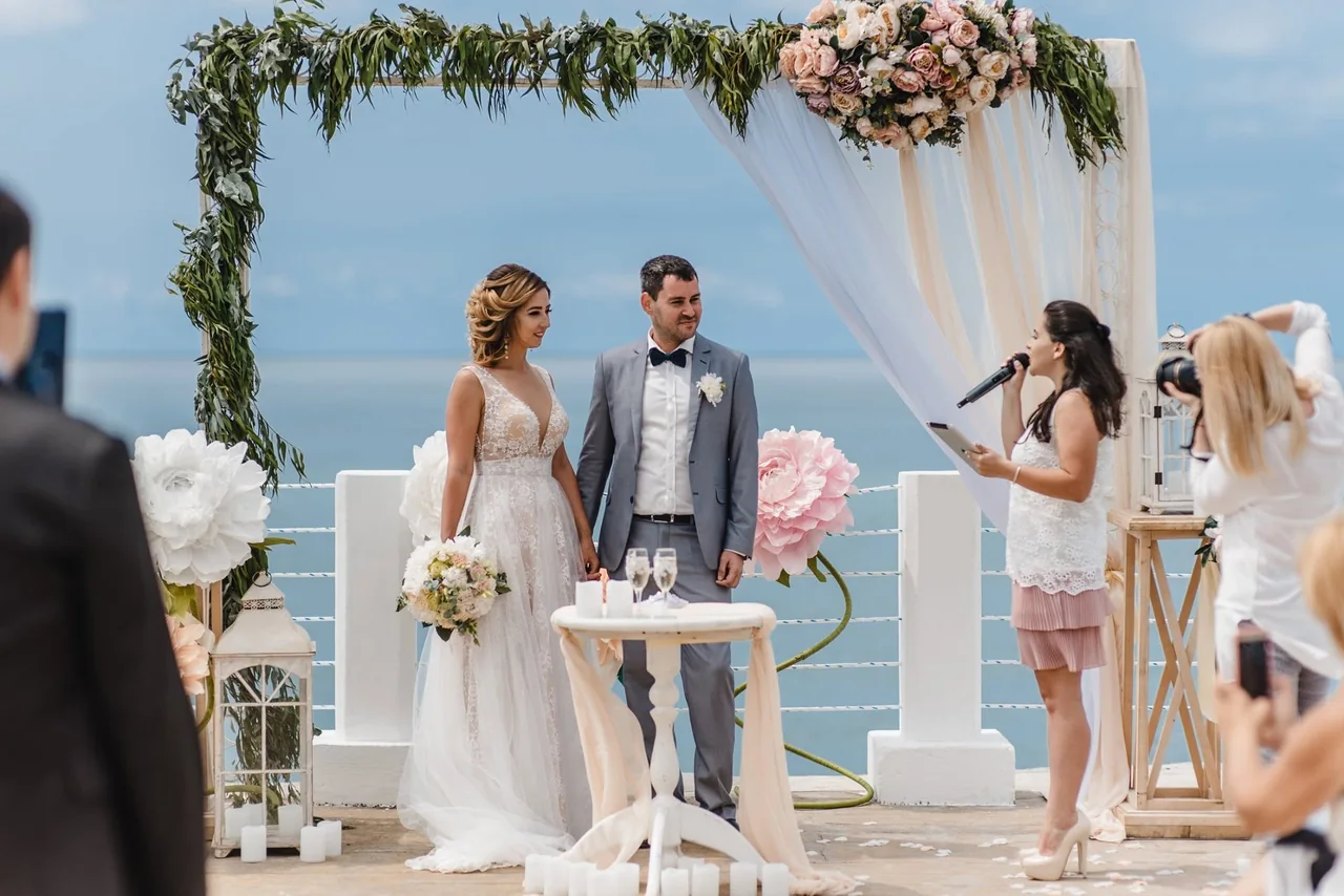 Ceremony on a white terrace above the sea: the couple at a flower arch and the celebrant with a microphone